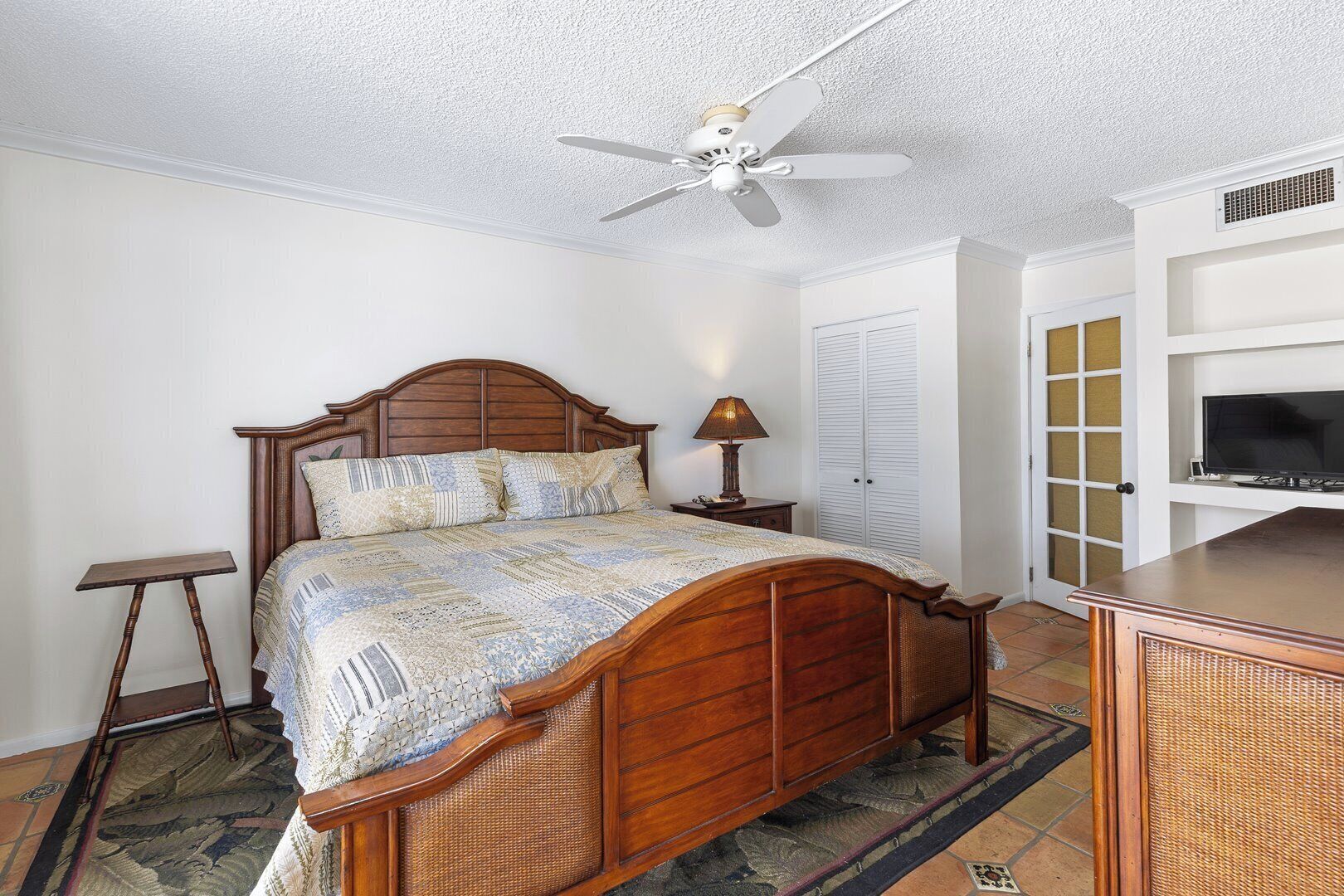 A bedroom with a wooden bed, a ceiling fan, a bedside table with a lamp, a TV on a stand, and a wardrobe.