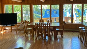 A wooden interior with a dining table and chairs, a sofa, and large windows offering a view of the outside.
