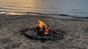 A beach scene with a campfire on the sand, a calm sea, and a sunset in the background.