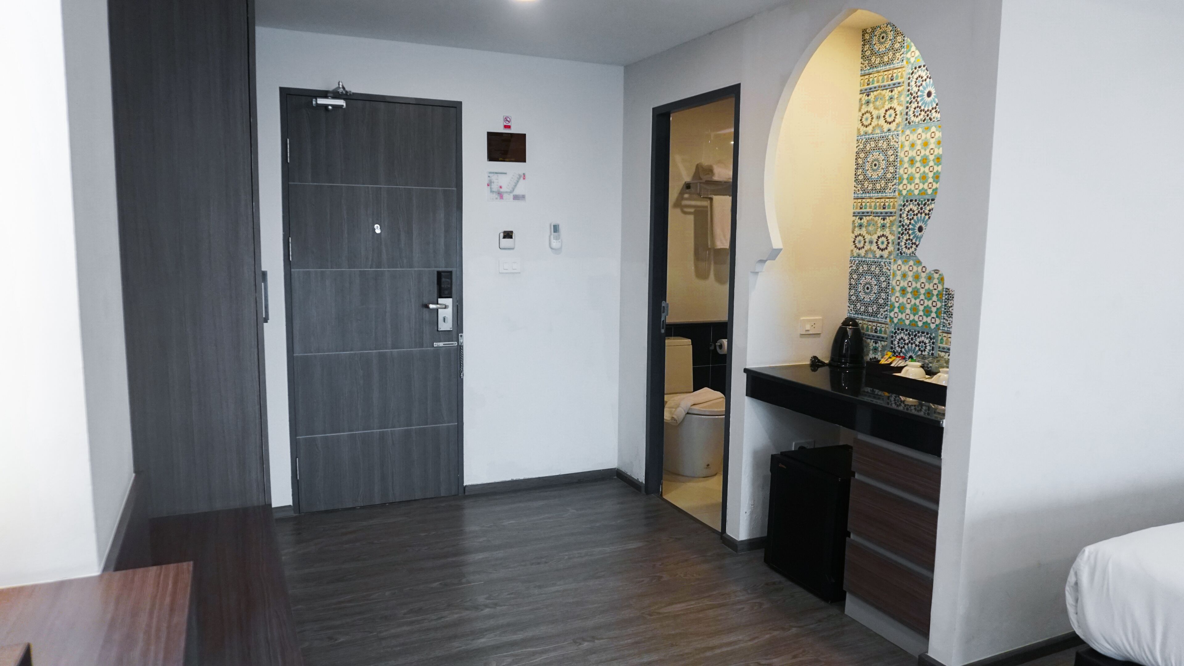 A hotel room with a dark wood floor, a door with a handle, a small window, and a tiled backsplash in the bathroom.