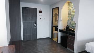 A hotel room with a dark wood floor, a door with a handle, a small window, and a tiled backsplash in the bathroom.