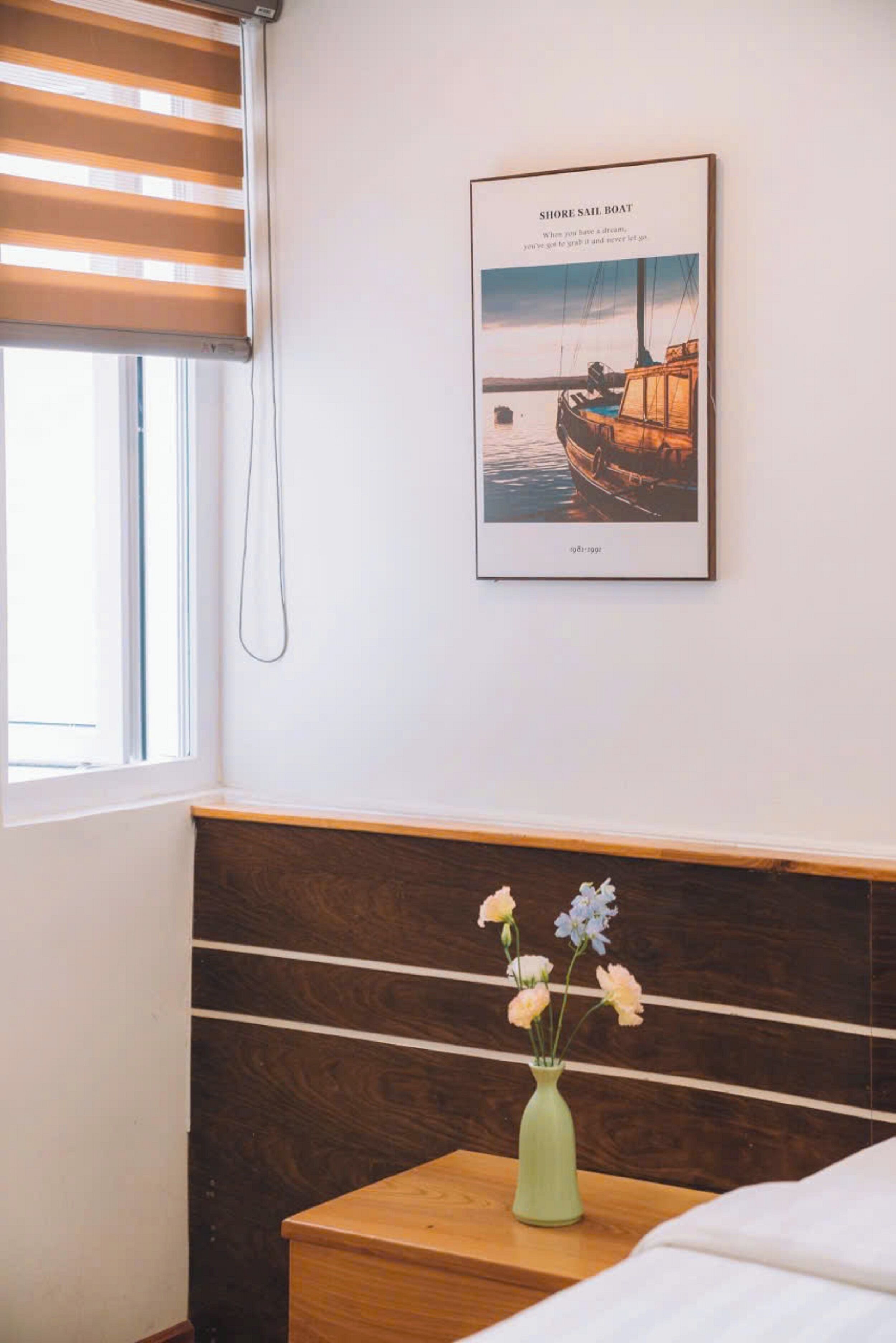 A bedroom with a bed, a nightstand with a vase of flowers, a framed picture of a sailboat, and a window with blinds.