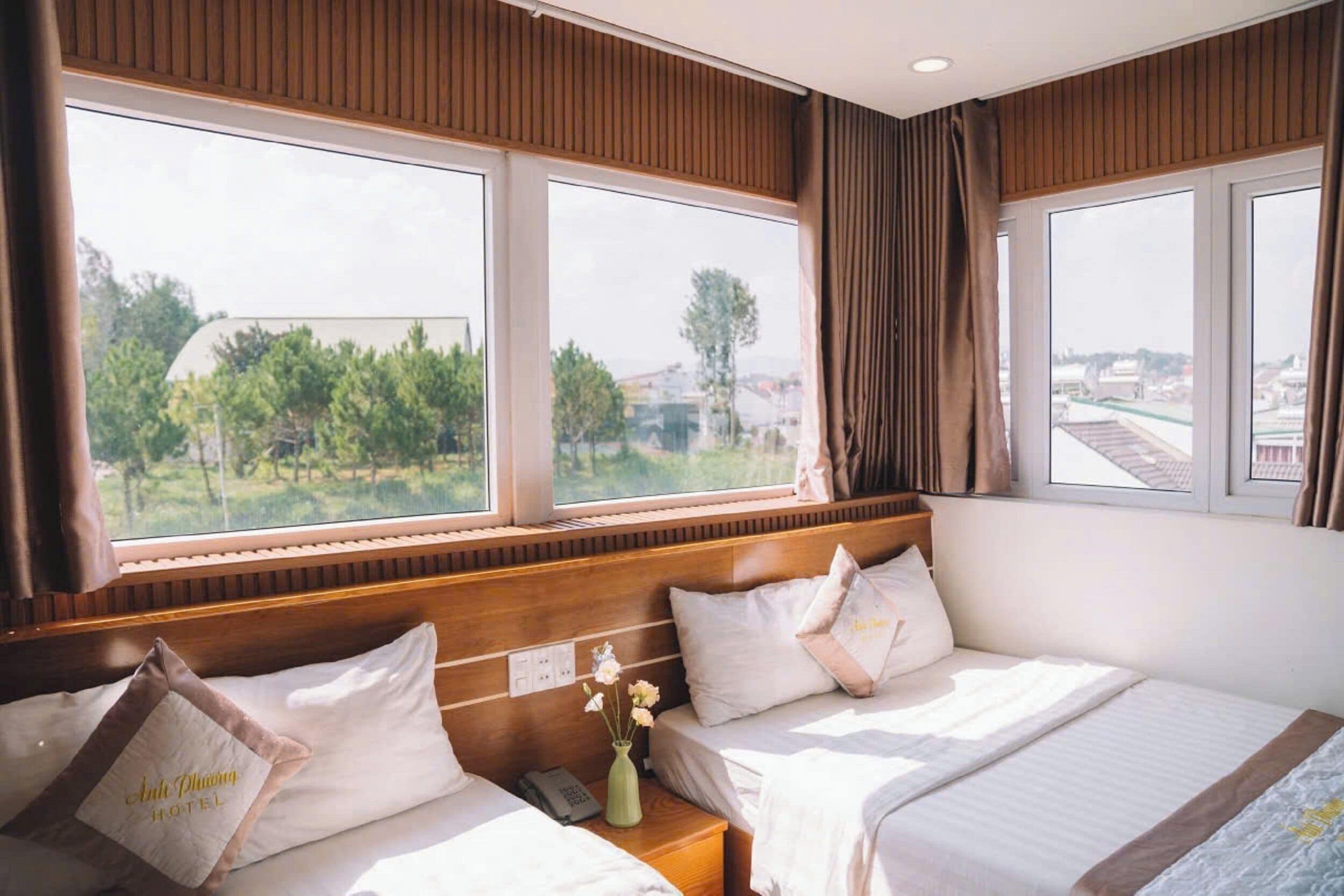 A hotel room with two beds, a wooden headboard, and large windows offering a view of greenery and buildings.