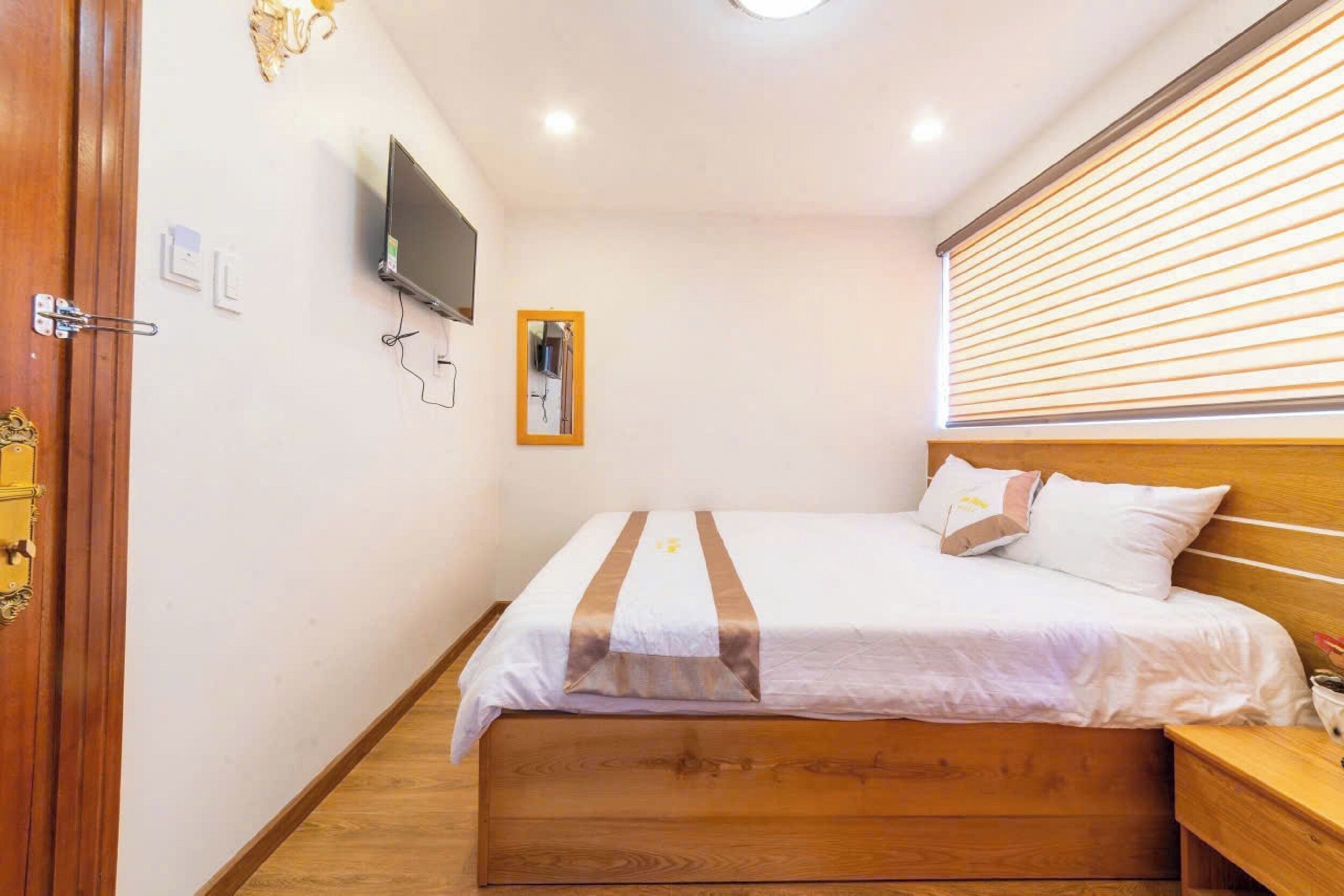 A bedroom with a wooden bed, a bedside table, a TV mounted on the wall, and a window with blinds.
