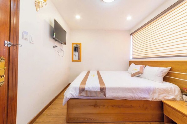 Une chambre à coucher avec un lit en bois, une table de chevet, un téléviseur fixé au mur et une fenêtre avec des stores.