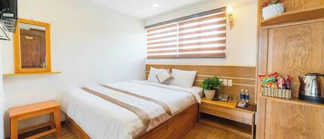 Habitación de hotel con cama de madera, mesita de noche con una planta, un pequeño banco, armario de madera y ventana con persianas.