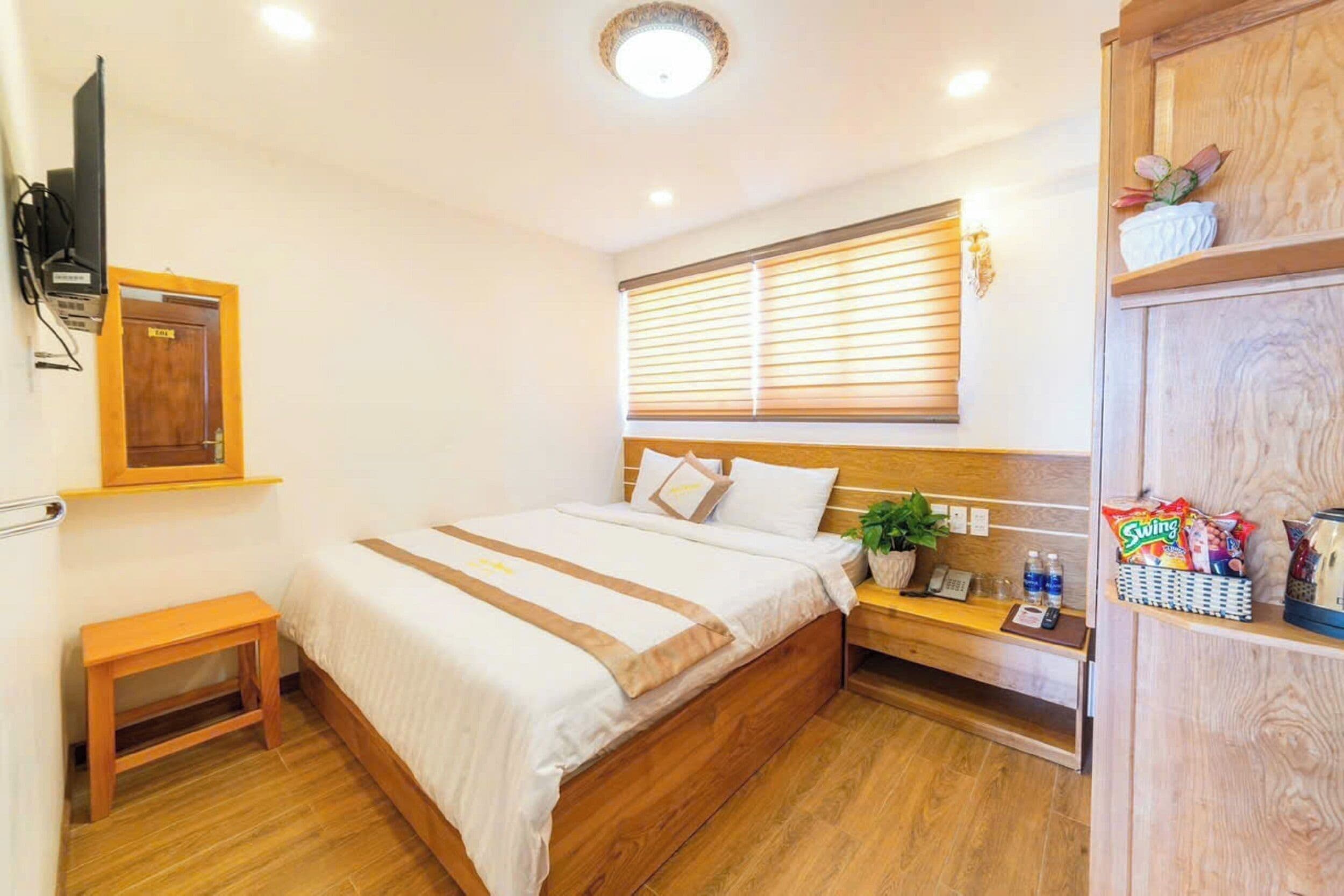 A hotel room with a bed, bedside table, a small plant, a shelf with snacks, and a TV mounted on the wall.