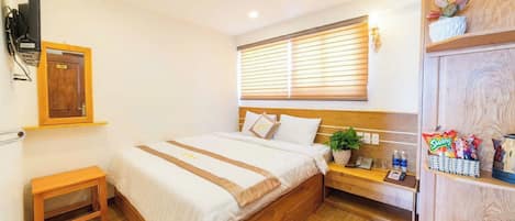 A hotel room with a bed, bedside table, a small plant, a shelf with snacks, and a TV mounted on the wall.