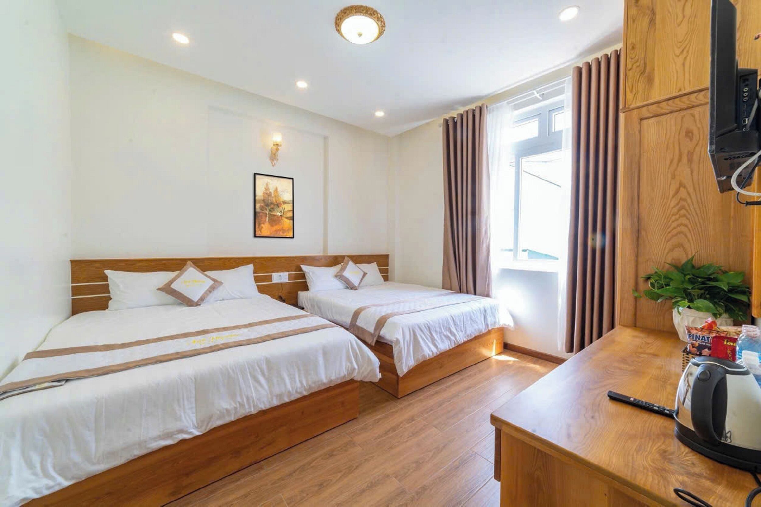 Une chambre d’hôtel avec deux lits, un plancher en bois, un tableau au mur, une fenêtre avec des rideaux et un bureau équipé d’une bouilloire et d’une télécommande.