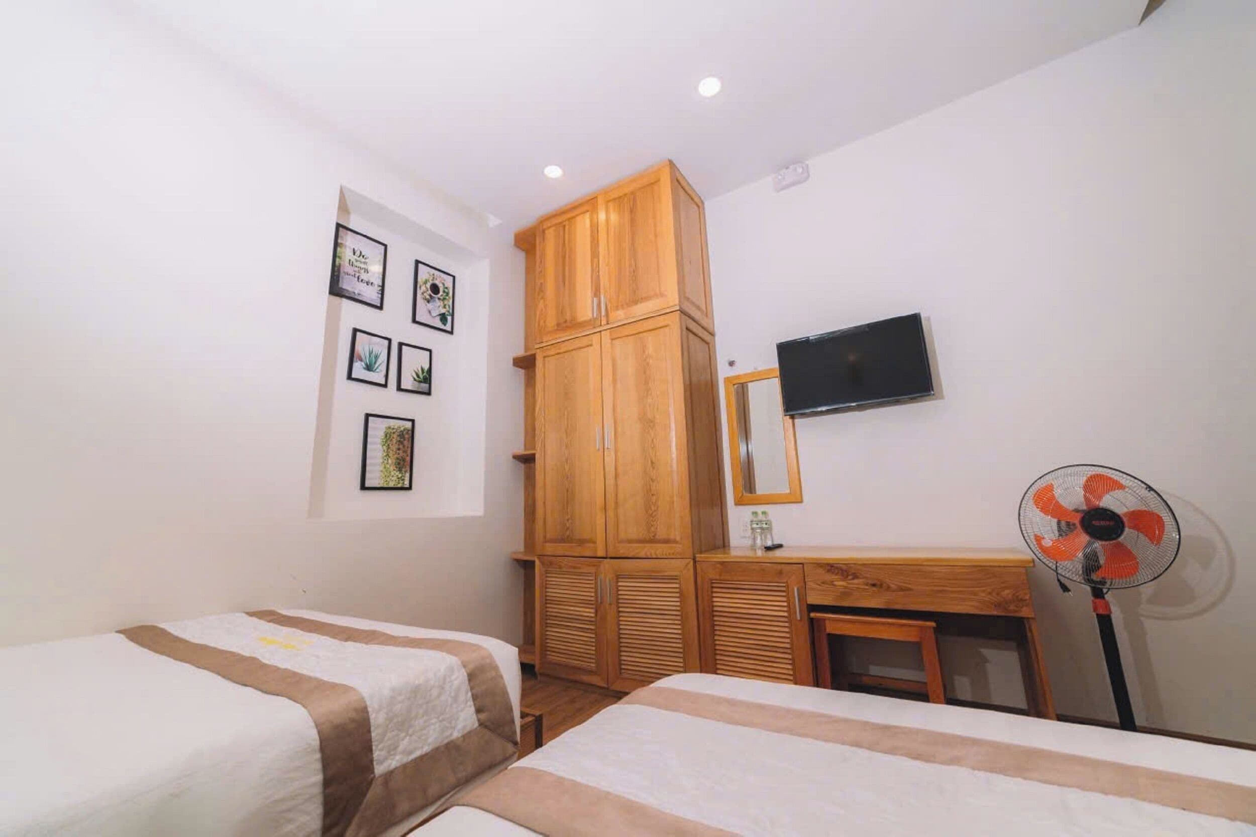 A bedroom with two beds, a wooden wardrobe, a desk with a mirror, and a standing fan.