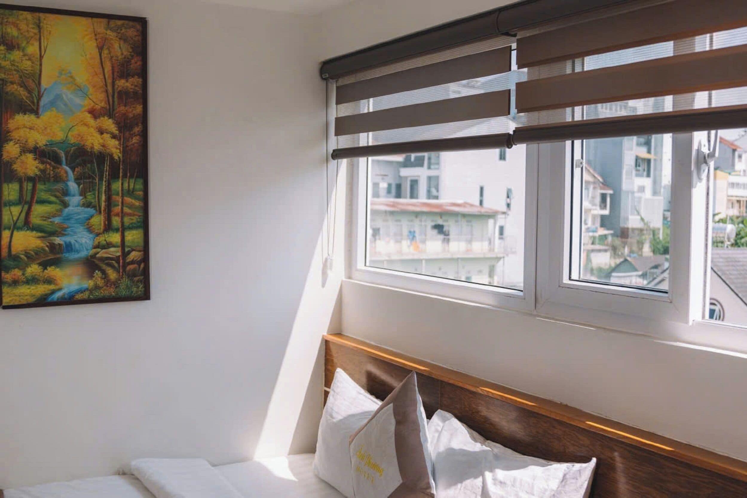 A hotel room with a bed, a painting of a forest scene, and a window with blinds.
