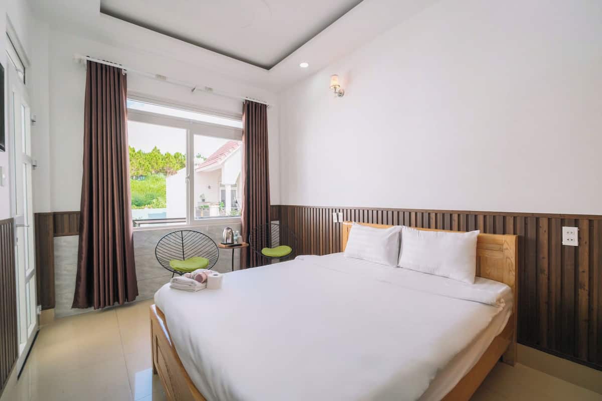 A hotel room with a large bed, a small table with a chair, a window with curtains, and a view of greenery outside.