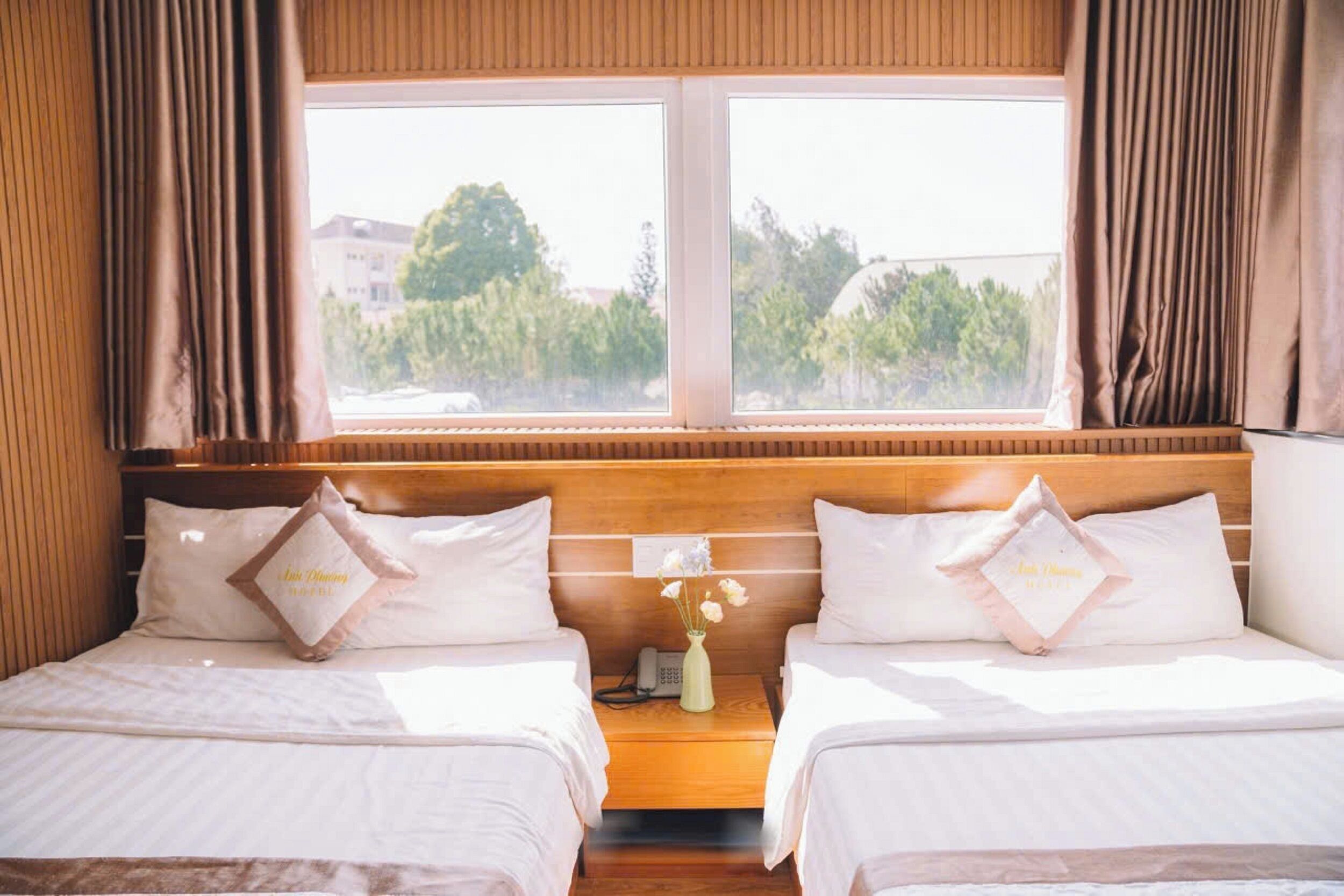 A hotel room with two beds, a wooden headboard, a nightstand with a vase of flowers, and a window with curtains.