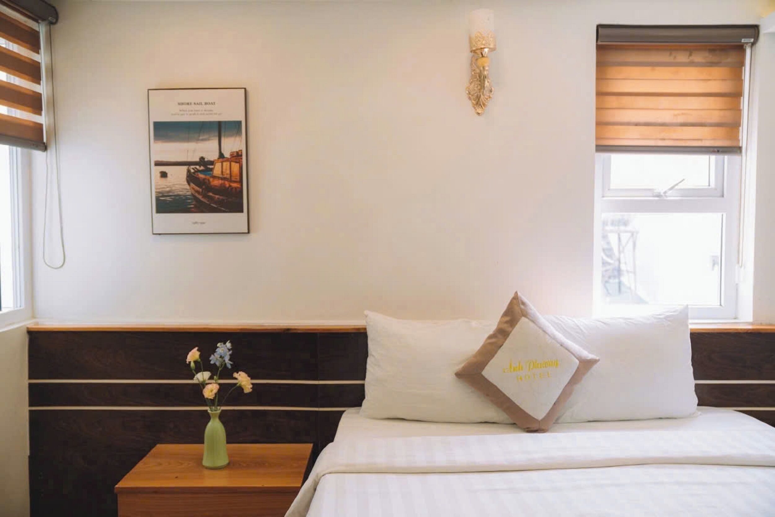 A hotel room with a bed, a nightstand with a vase of flowers, a framed picture on the wall, and a window with blinds.