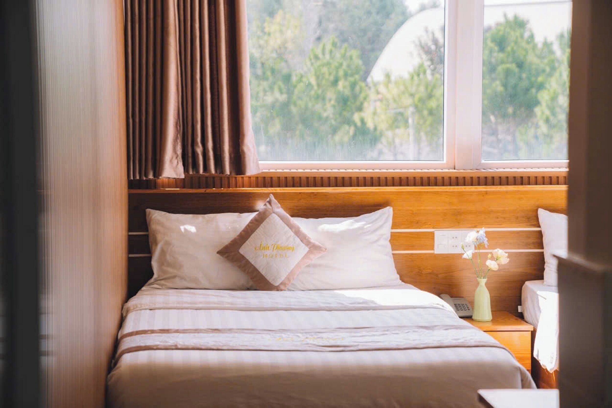 Une chambre d’hôtel avec un lit, une table de chevet, une fenêtre avec des rideaux et une vue sur la verdure.