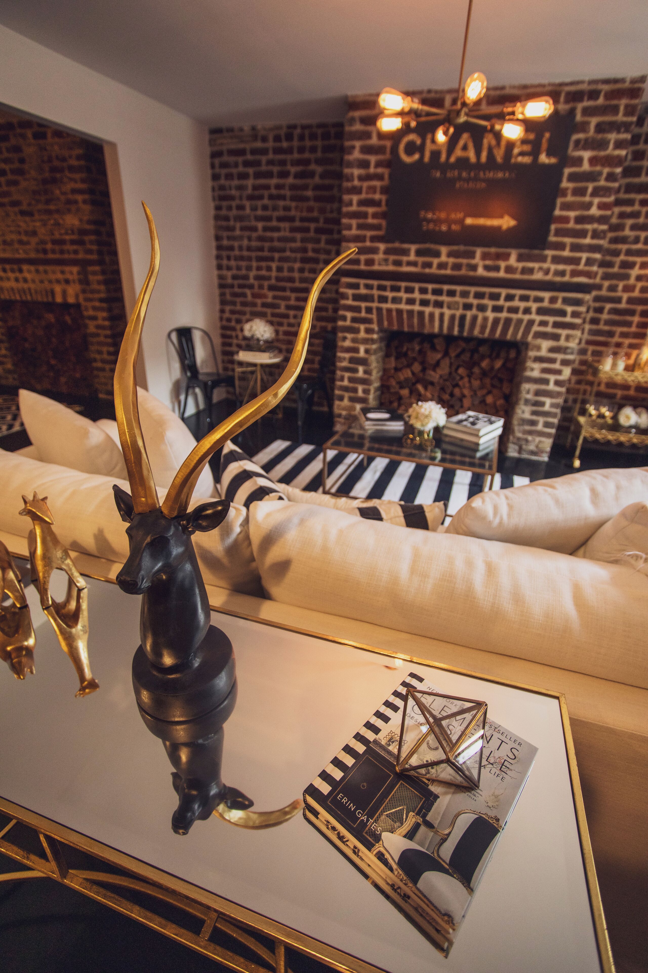 A modern living room with a sofa, a coffee table with a magazine, and a decorative antelope head. A brick wall with a 'CHANEL' sign is in the background.