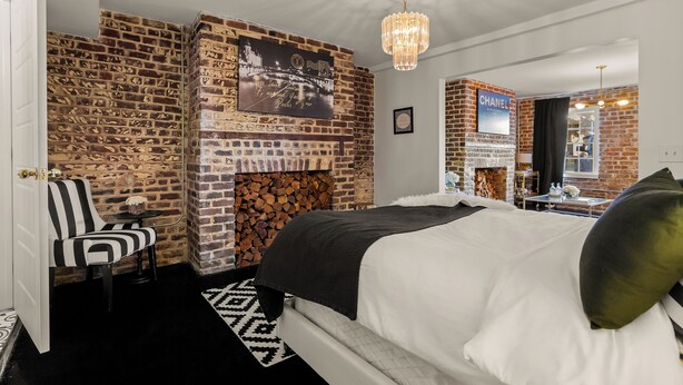 A bedroom with a brick wall, a fireplace, a bed with white bedding, a black and white striped chair, and a chandelier. - Luxury Townhouse Chic Flat in the Historic Center! (savannah)