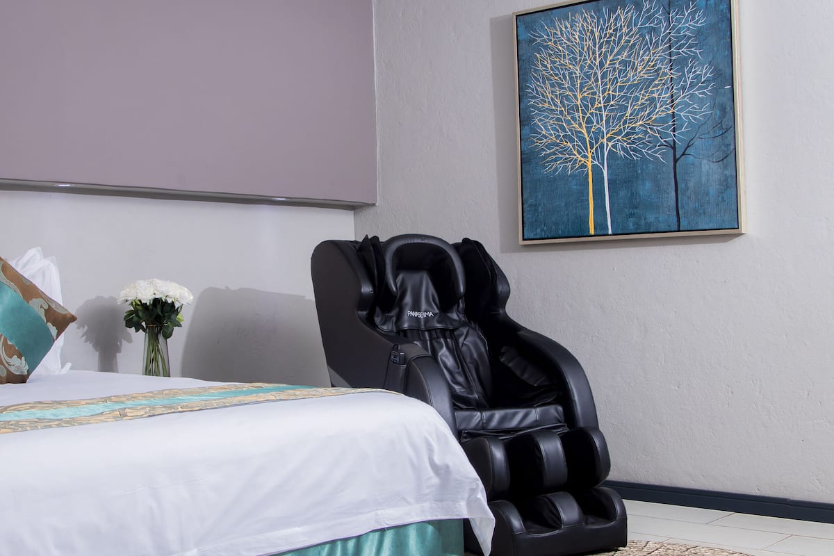 A bedroom with a bed, pillows, a massage chair, a vase with flowers, and a painting on the wall.