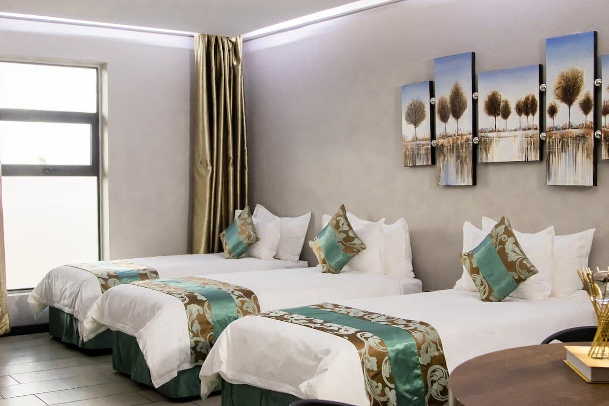 A hotel room with two beds, a large window with curtains, a wall with four paintings, and a small table with a book and a decorative item.