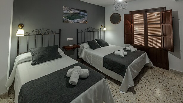 Habitación de hotel con dos camas, un cuadro en la pared, un espejo redondo y una ventana con contraventanas de madera.