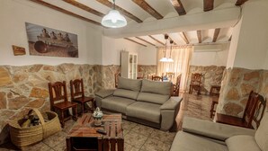 Una sala de estar acogedora con pared de piedra, un sofá color beige, sillas de madera, una mesita de centro y una lámpara colgante.