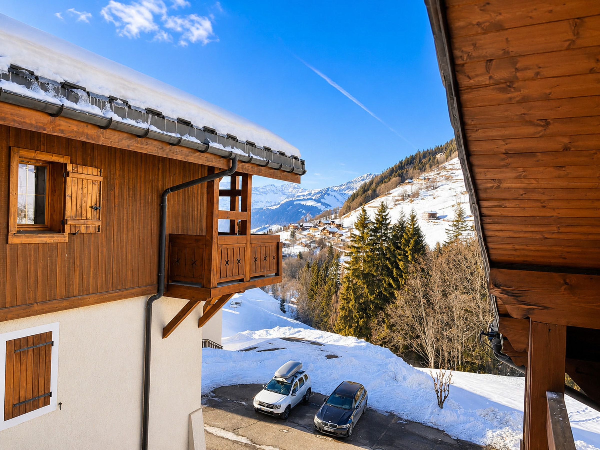 Een houten chalet met een balkon met uitzicht op een besneeerd berglandschap en geparkeerde auto's.