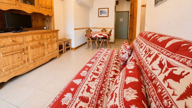 Una habitación con un armario de madera, un televisor, un sofá estampado, una mesa de comedor con sillas y un cuadro en la pared.