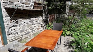 Une table et des chaises en bois, placées contre un mur de pierre, le tout entouré de verdure.