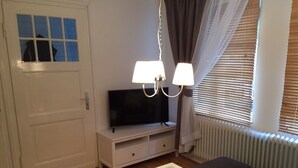 Une pièce avec un téléviseur posé sur un meuble blanc, une table avec des bougies, une chaise et une fenêtre avec des stores.