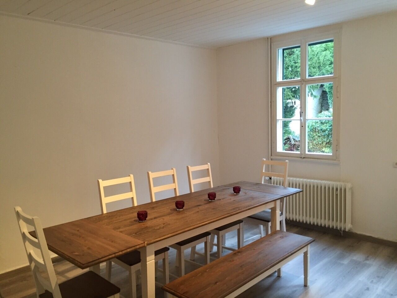 Zona pranzo con tavolo in legno, sedie bianche e una finestra vista su vegetazione.