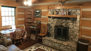 A cozy cabin interior with a stone fireplace, wooden walls, a bookshelf, and a dining area.