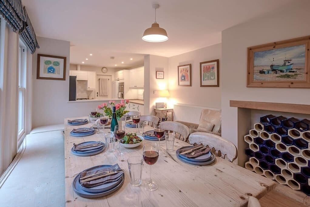 Une salle à manger avec une longue table dressée pour un repas, entourée de chaises. On y trouve une bouteille de vin et des verres sur la table, ainsi qu’un vase rempli de fleurs. La pièce dégage une atmosphère chaleureuse, avec une lampe et des œuvres d’art accrochées aux murs.