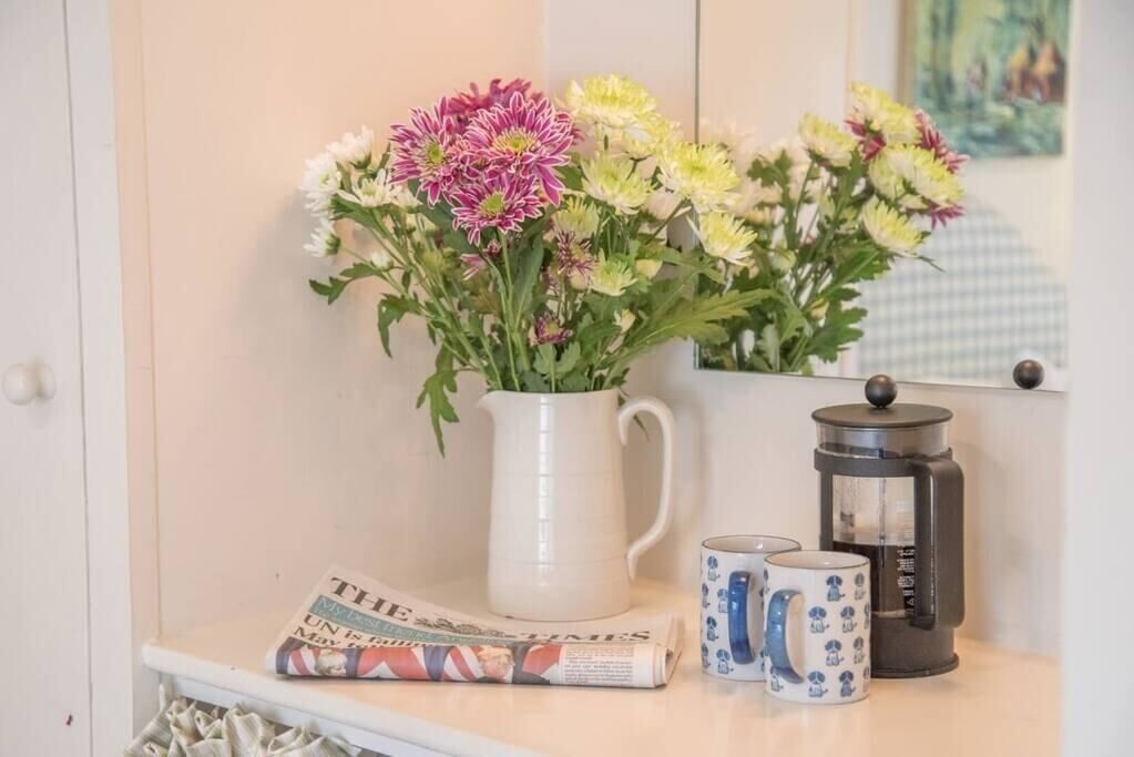 Uma caneca branca, um jornal e um jarro branco com flores em uma estante.