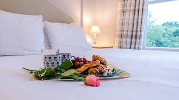 A neatly made bed with white linens, a plate of croissants and grapes, a cup with a blue pattern, and a rose on the bed.