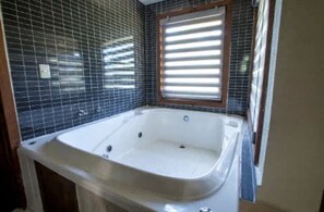 Ein Badezimmer mit einer großen Badewanne, gefliesten Wänden und einem Fenster mit Jalousien.