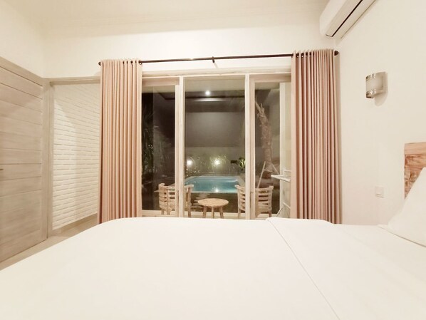 Un dormitorio con una cama grande, un vidrio corredero que da a una zona de piscina y una pequeña mesa con sillas.