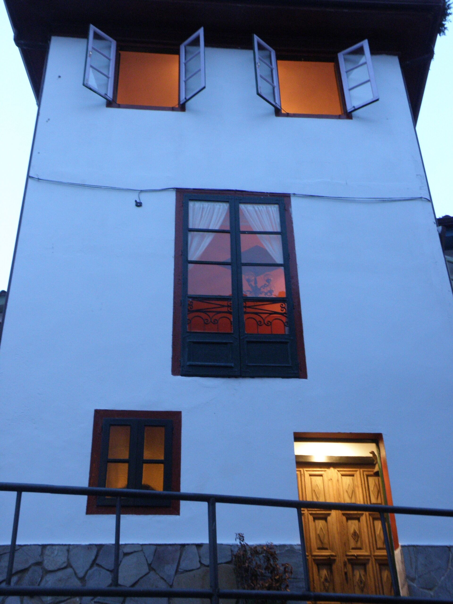 Un bâtiment à deux étages, doté d’une façade blanche, d’une fenêtre centrale avec des volets noirs et d’une porte en bois située en bas.