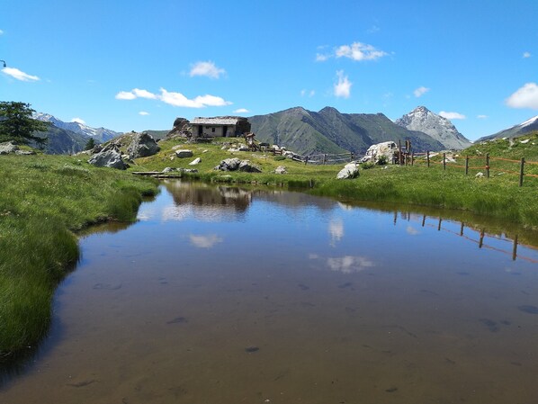 Una cabaña de montaña con un estanque en primer plano, rodeada de terreno rocoso y montañas.