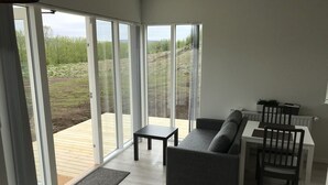 A modern living room with a sofa, a small table, and a dining area with chairs. There are large sliding glass doors leading to an outdoor deck.