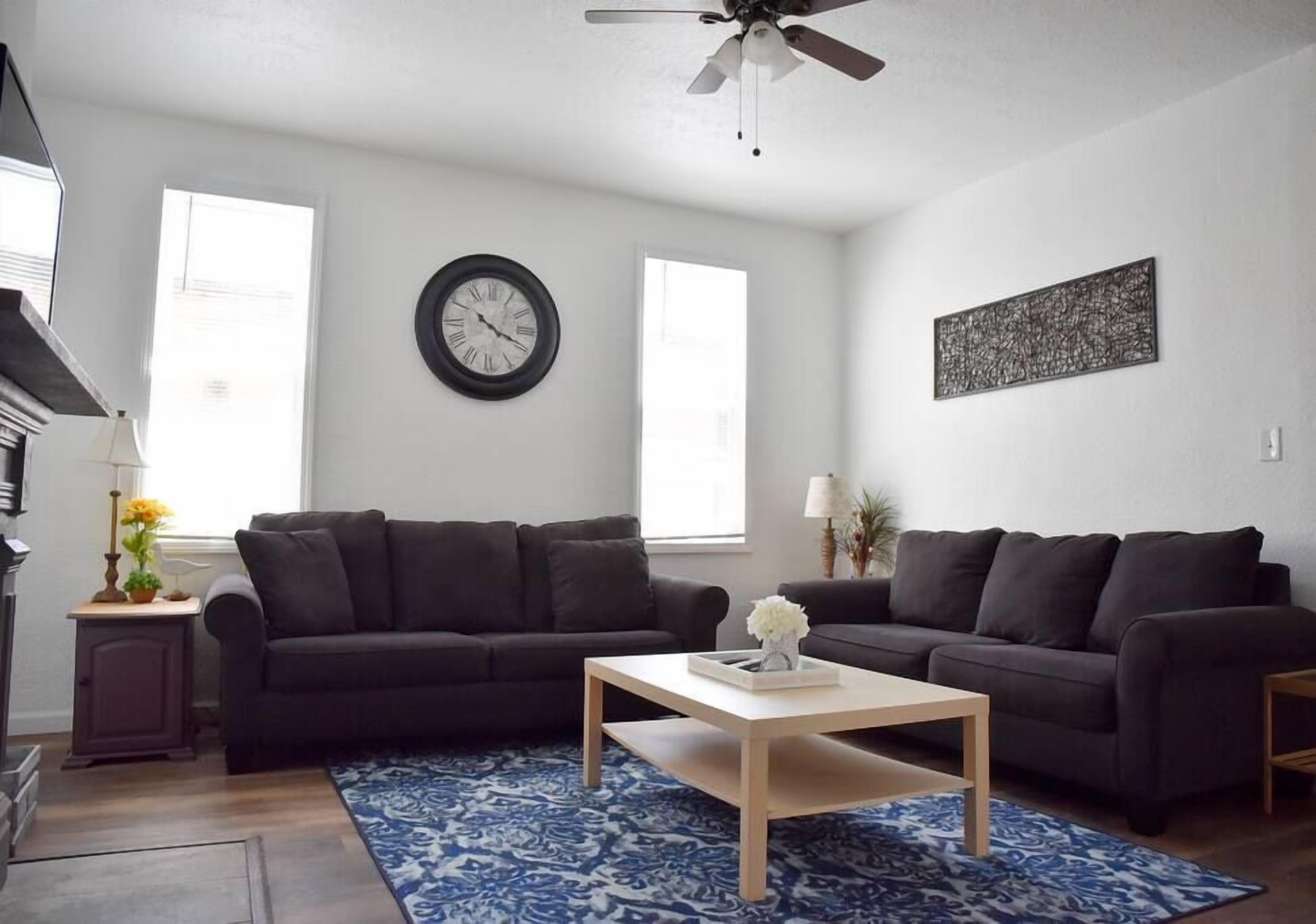 Ein Wohnzimmer mit zwei dunklen Sofas, einem Holztisch, einem blauen Teppich mit Muster, einer Wanduhr und einem Deckenventilator.