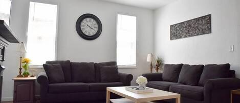 Ein Wohnzimmer mit zwei dunklen Sofas, einem Holztisch, einem blauen Teppich mit Muster, einer Wanduhr und einem Deckenventilator.