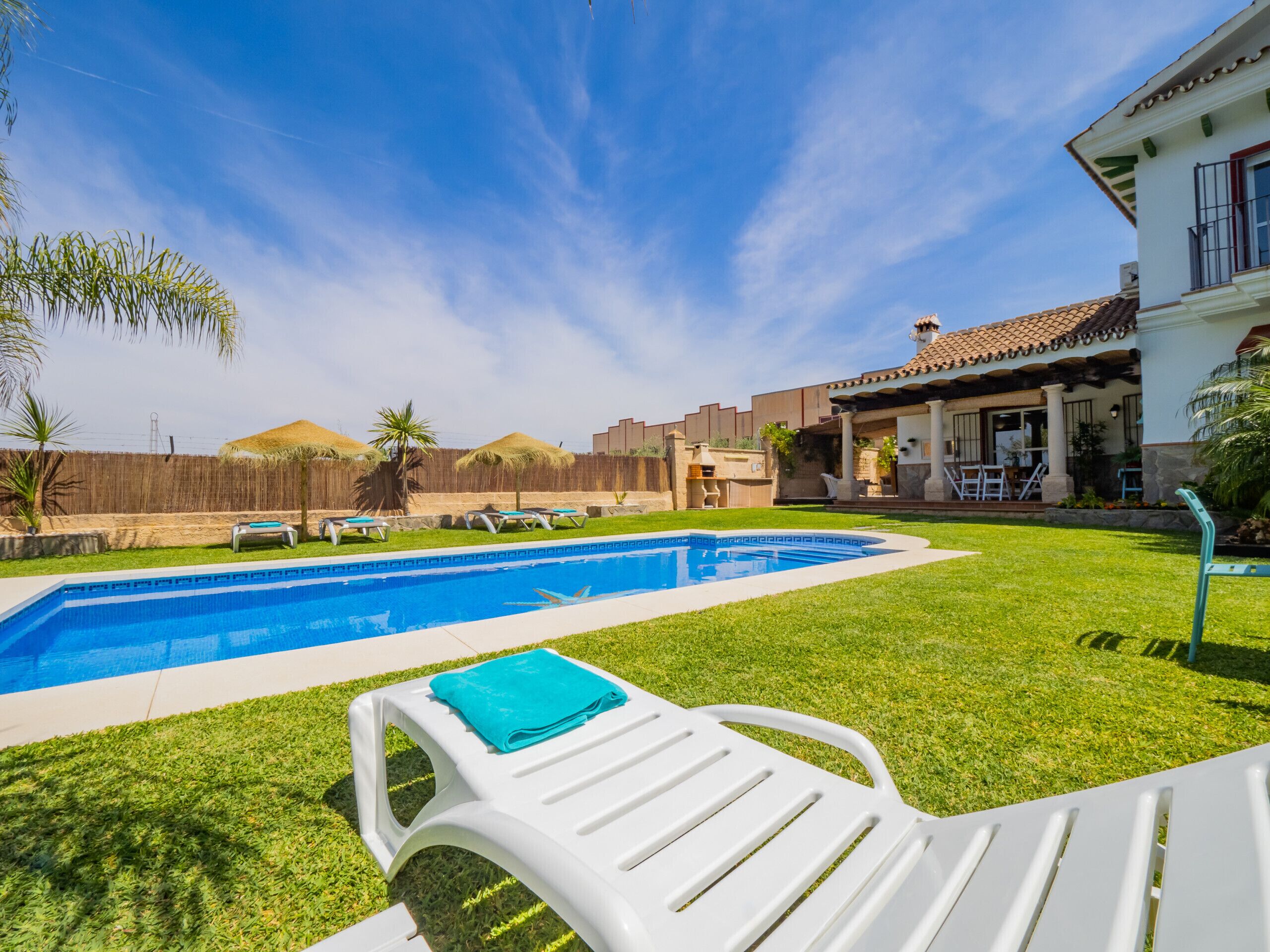 Une piscine, avec une chaise longue blanche et une serviette bleue, entourée d’une pelouse bien entretenue et d’une maison dont le toit est recouvert de tuiles.
