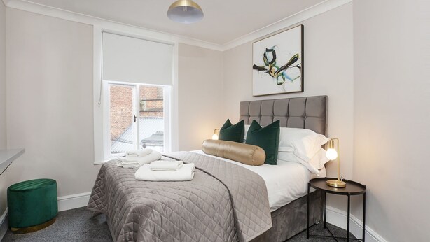 A modern bedroom with a large bed, a window with a view of a brick building, a green ottoman, and a framed abstract painting on the wall. - Sleeps 6 guests  in 2 bedrooms (York)