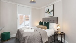 Un dormitorio moderno con una cama grande, una ventana con vista a un edificio de ladrillo, un otoman verde y un cuadro abstracto enmarcado en la pared.