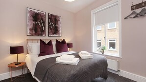 A neatly made bed with a grey blanket, two purple pillows, and a white duvet. There are two abstract paintings on the wall, a window with a view of a brick building, and a small plant on the windowsill. - Sleeps 6 guests  in 2 bedrooms (York)