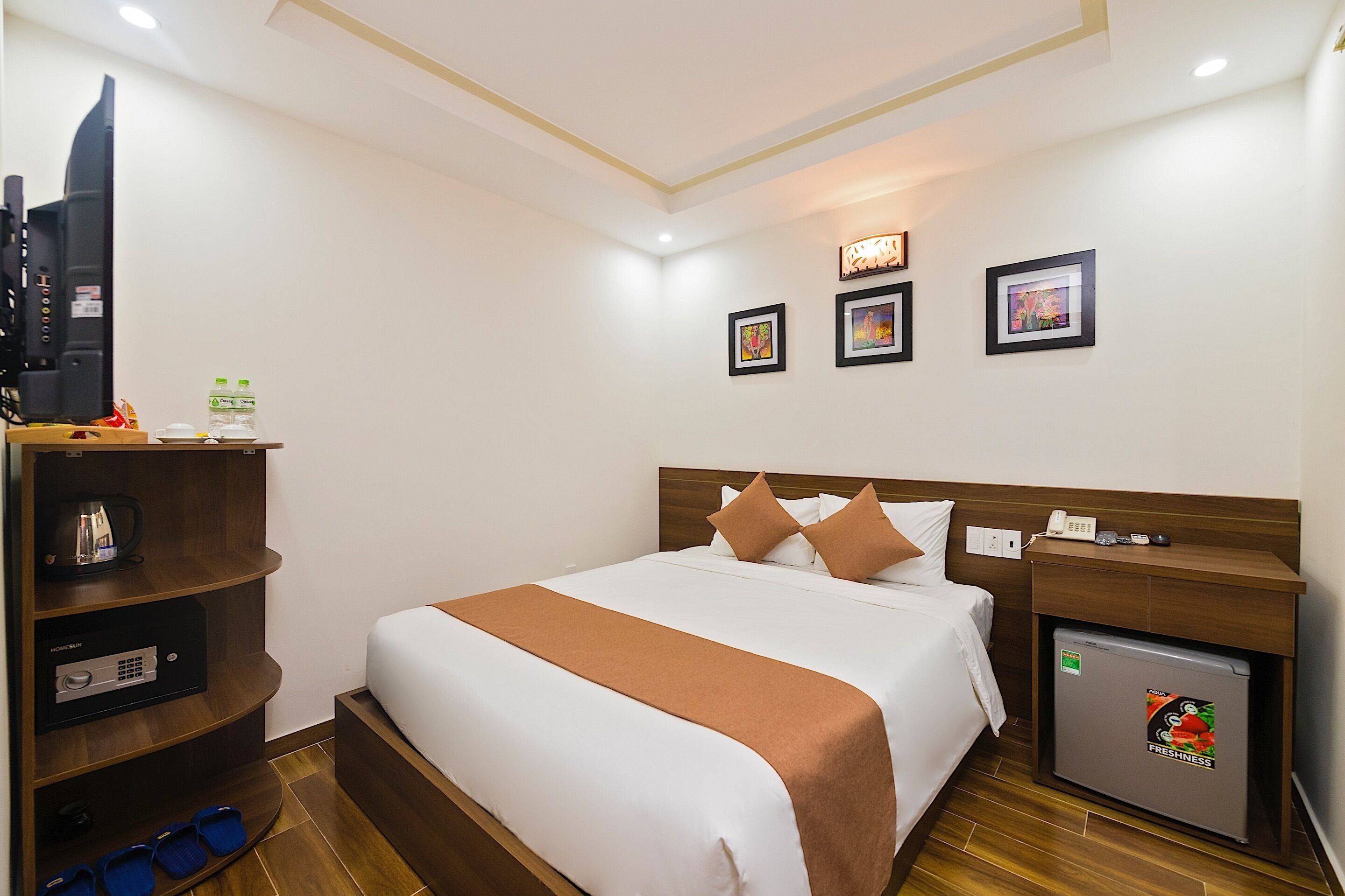 A hotel room with a bed, a TV, a mini-fridge, a wooden shelf with a kettle, and a small table with a phone and a cup.