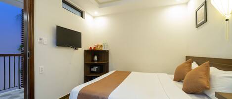 A hotel room with a bed, a TV mounted on the wall, a bedside table, a nightstand, a shelf with toiletries, and a window with a view of the outside.