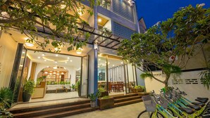 A modern hotel entrance with a glass facade, outdoor seating, and a bike rental station. - Brother Villa Hoi An (Da Nang)