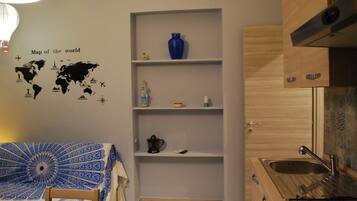 A compact kitchen with a gas stove, a sink, and a blue vase on a shelf. There's a world map on the wall and a bed with a blue and white patterned bedspread.