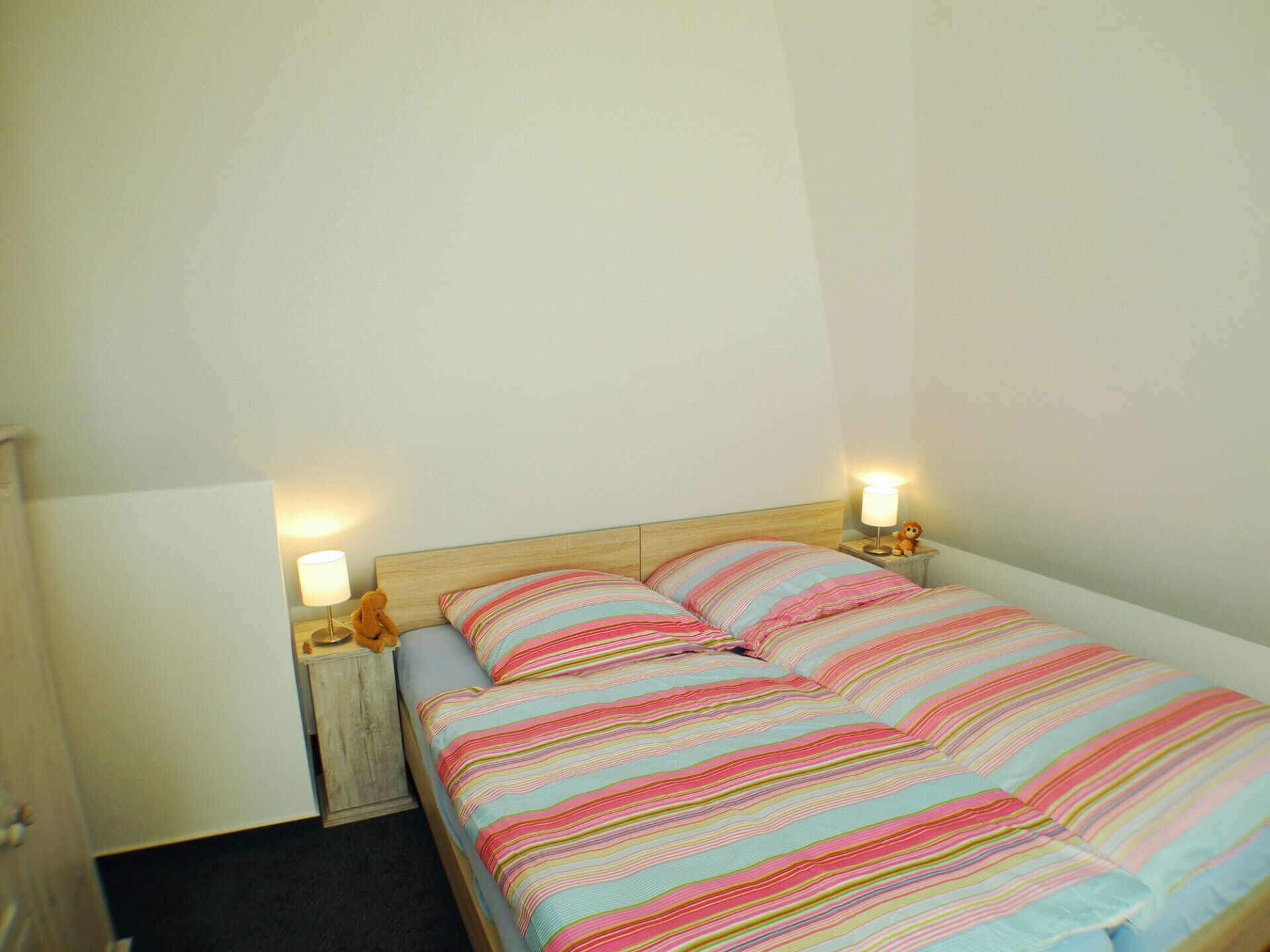 Un lit simple avec une couvre-lit à rayures, deux lampes de chevet et une table de chevet sur laquelle est posé un ours en peluche.